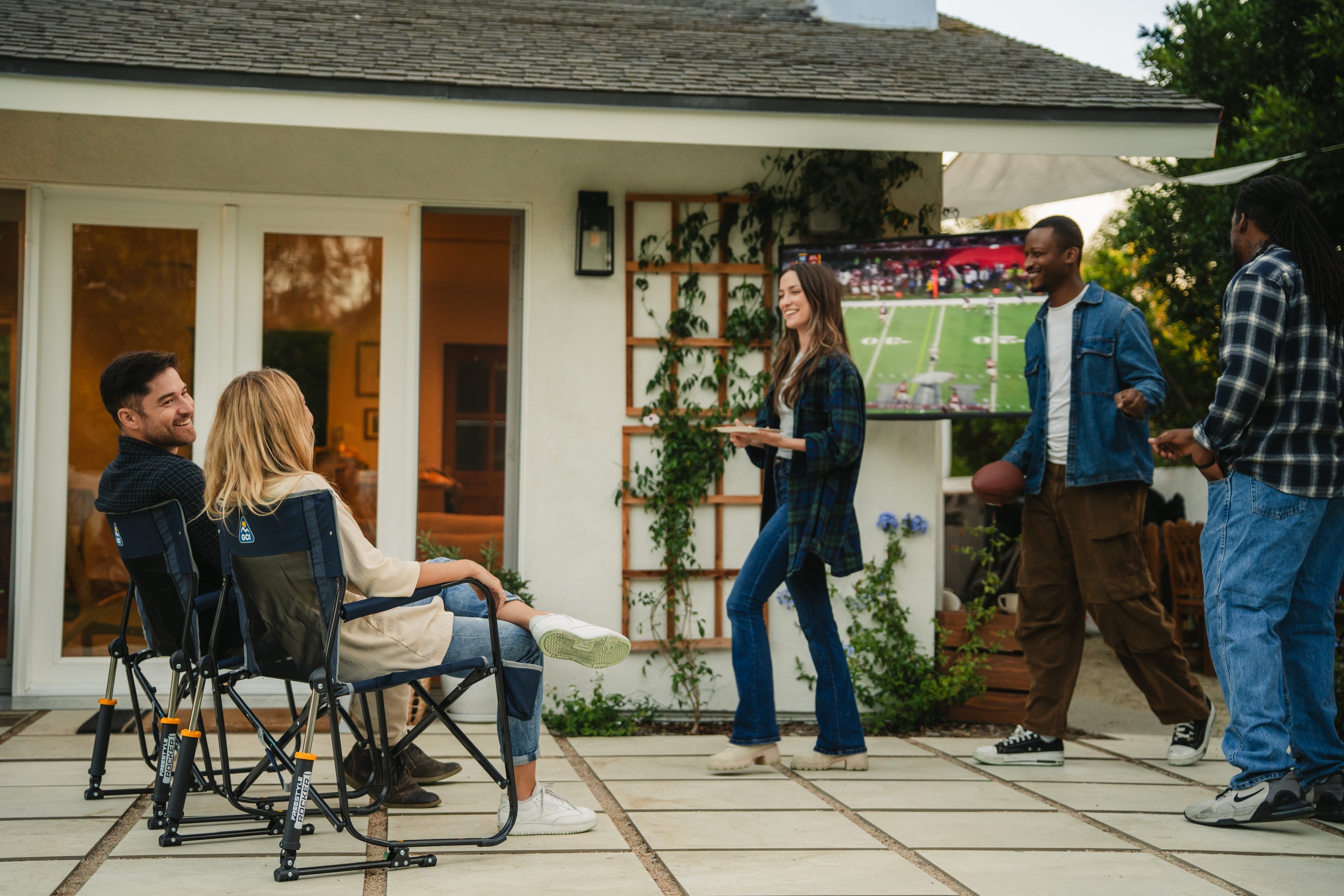 people at a backyard football watch party - two watching tv in GCI rocker, one carrying food, one holding football, and one standing